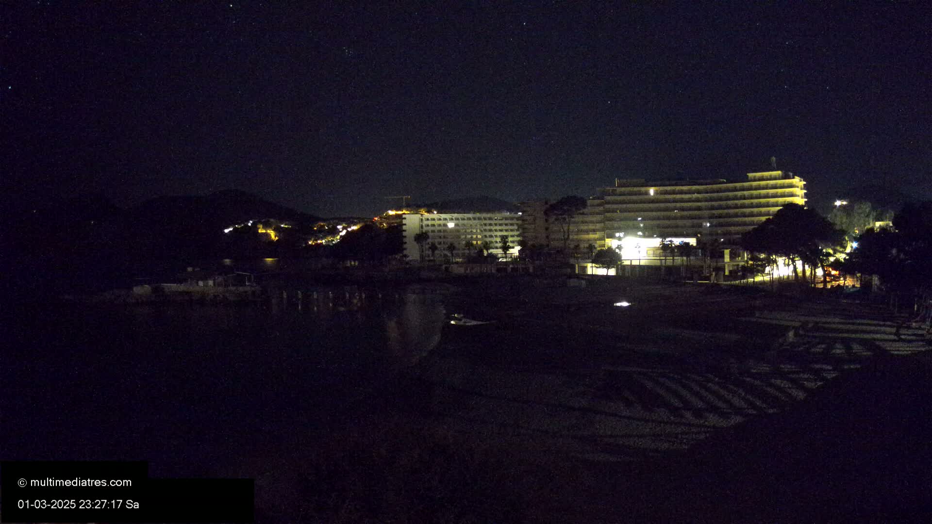 Under a starry night sky, illuminated hotels and a beach are visible near a dark mountainside.