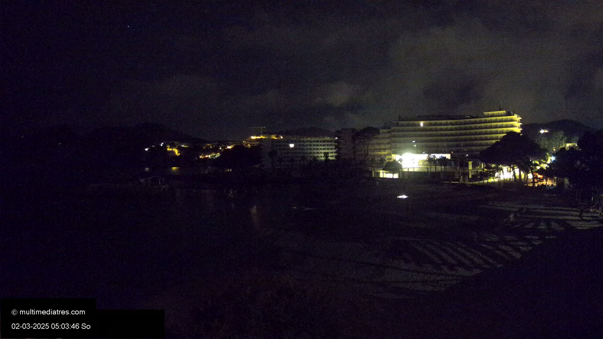 At night, under a partly cloudy sky, a large illuminated hotel stands near a dark beach and a dimly lit hillside.