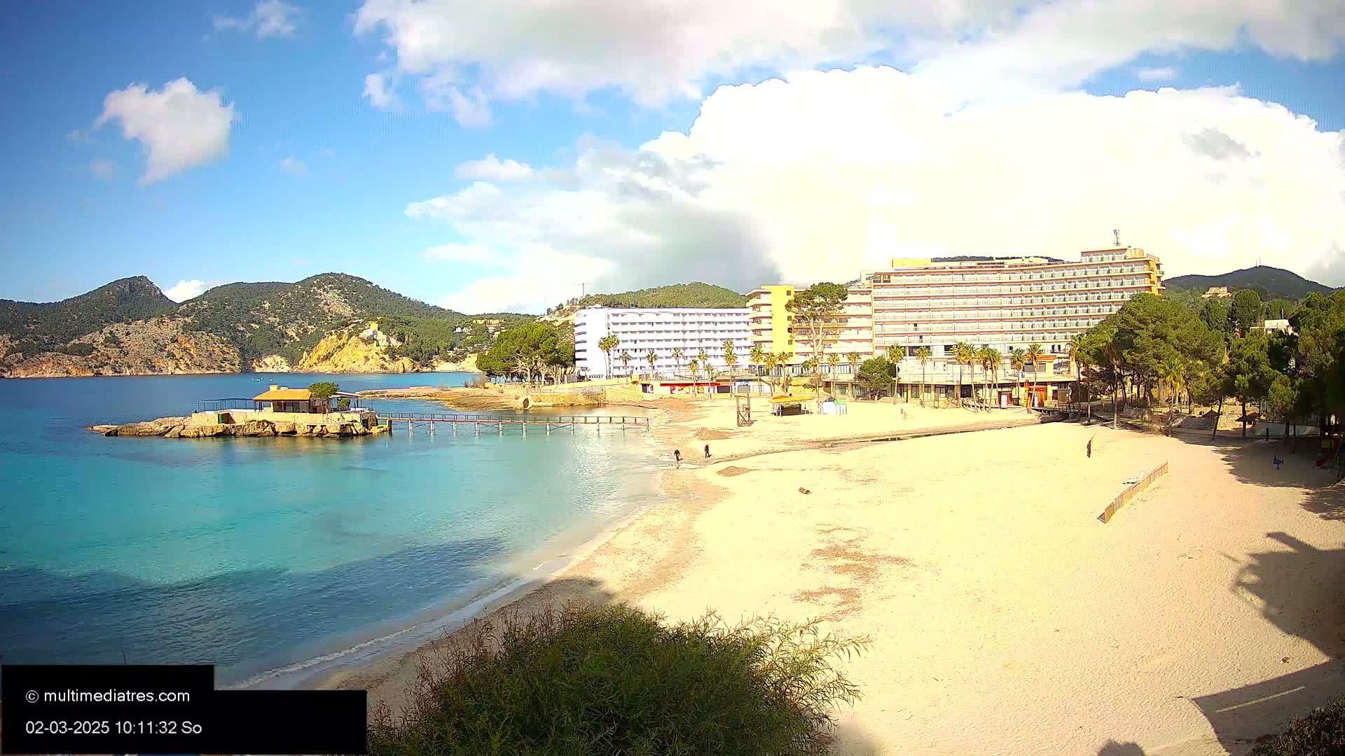 A sandy beach with turquoise water, a small building on a rock outcropping, a large hotel, and green hills under a partly cloudy sky.