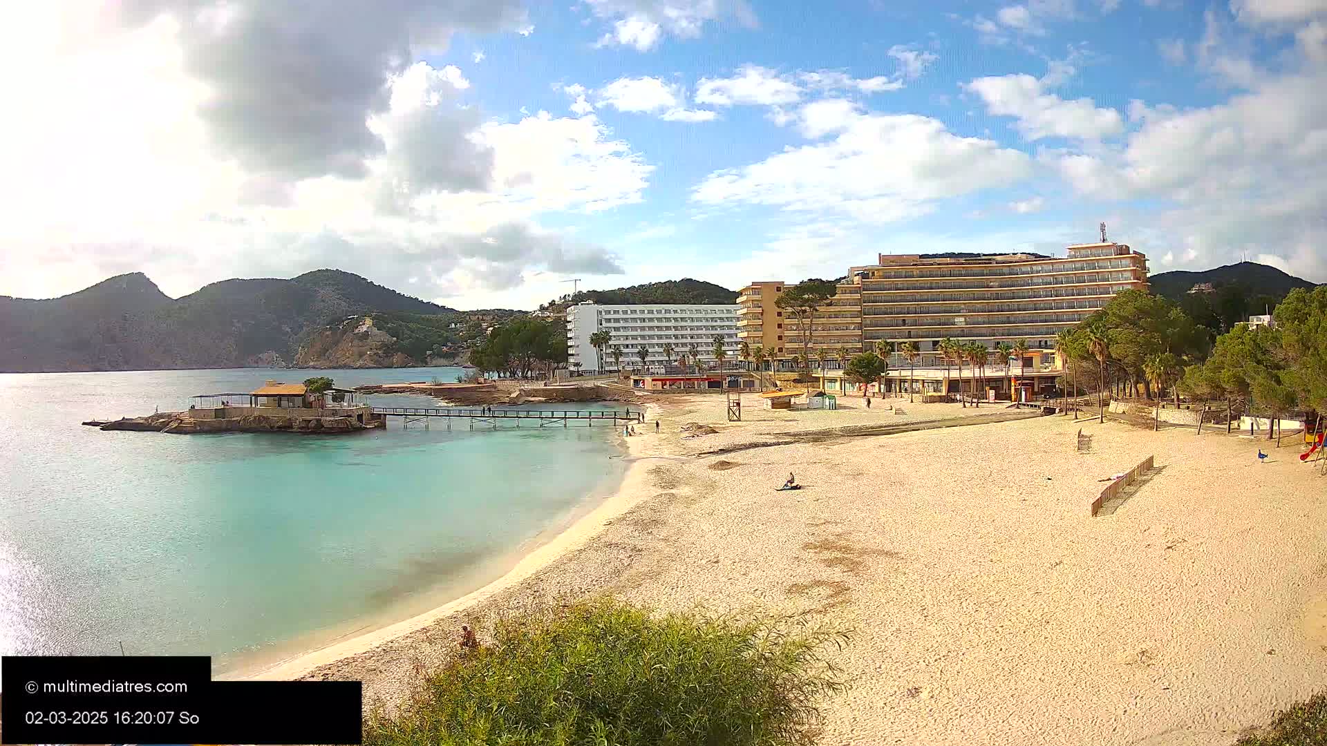 A sandy beach with calm, turquoise water and several hotels is seen under partly cloudy skies, with mountains in the background.