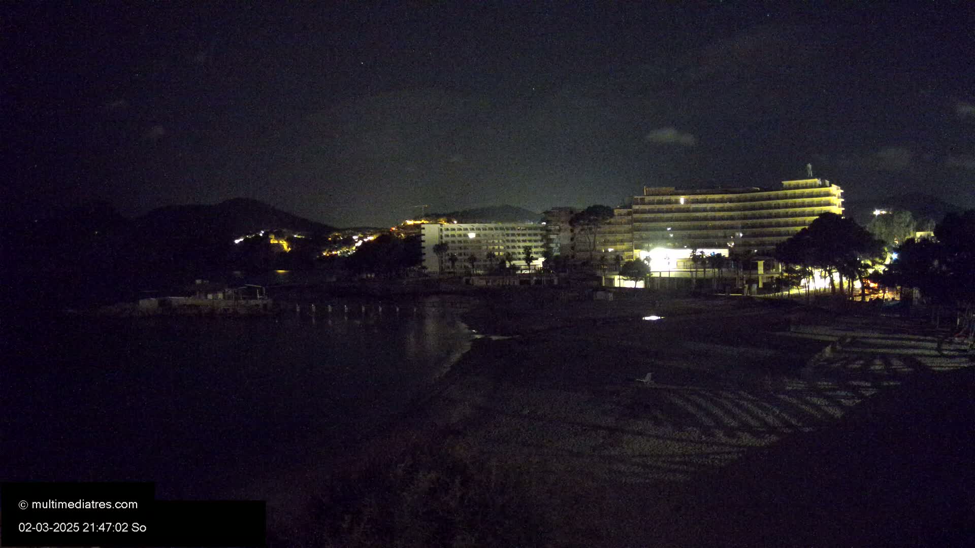 A nighttime view of a beach with a large hotel and other buildings illuminated against a dark, starlit sky.
