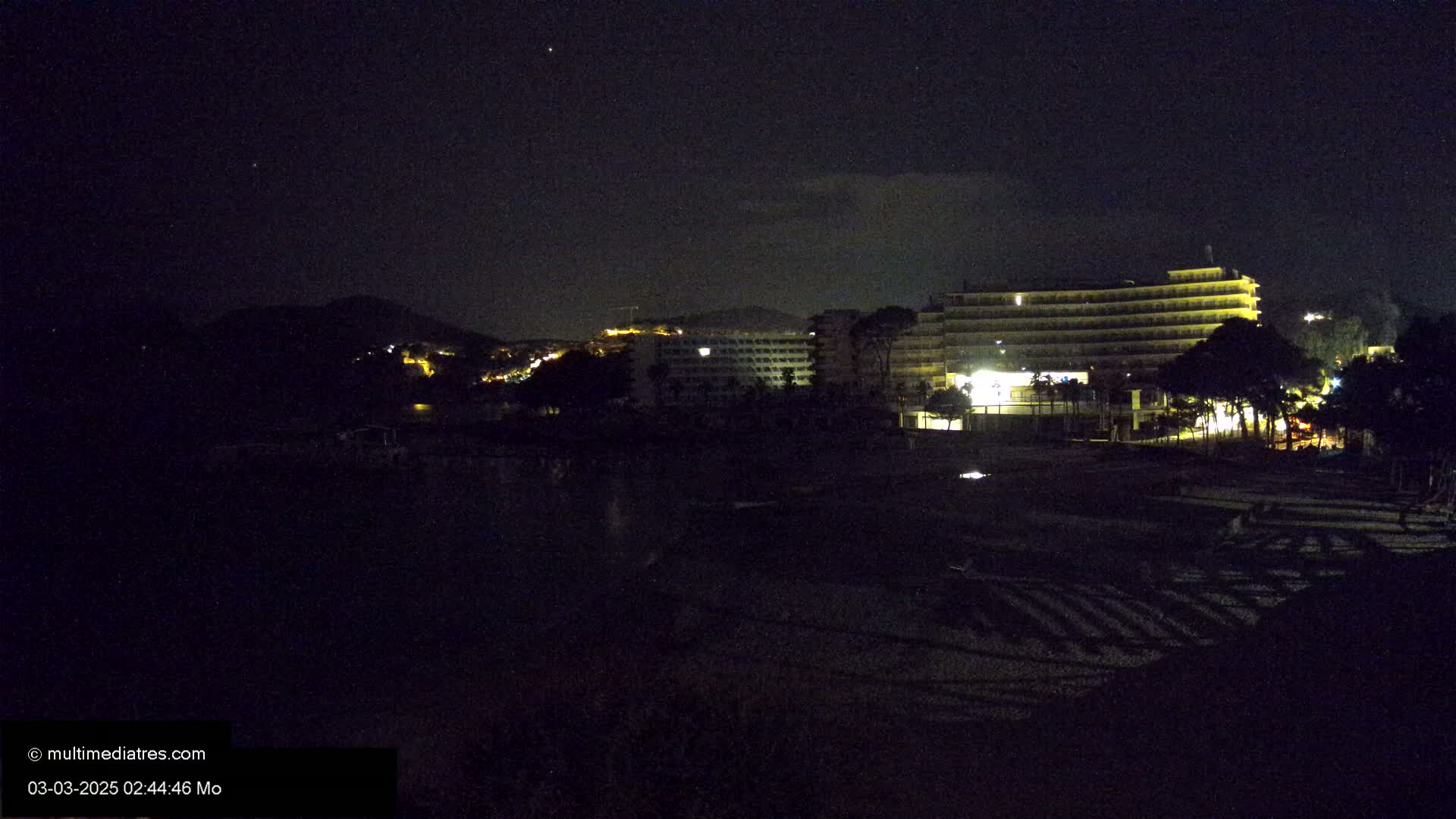 A dark beach at night, under a mostly clear sky, with several illuminated hotels and a hill in the background.