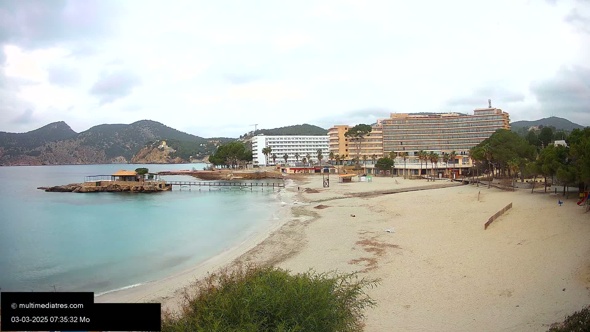 A sandy beach with turquoise water and two hotels is visible, backed by mountains under an overcast sky.
