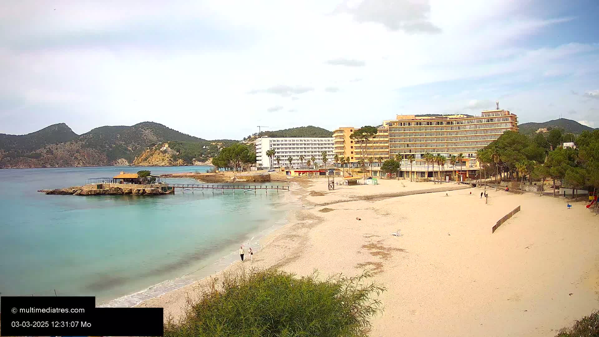A sandy beach with turquoise water stretches out before a row of hotels and lush green hills under a mostly cloudy sky.