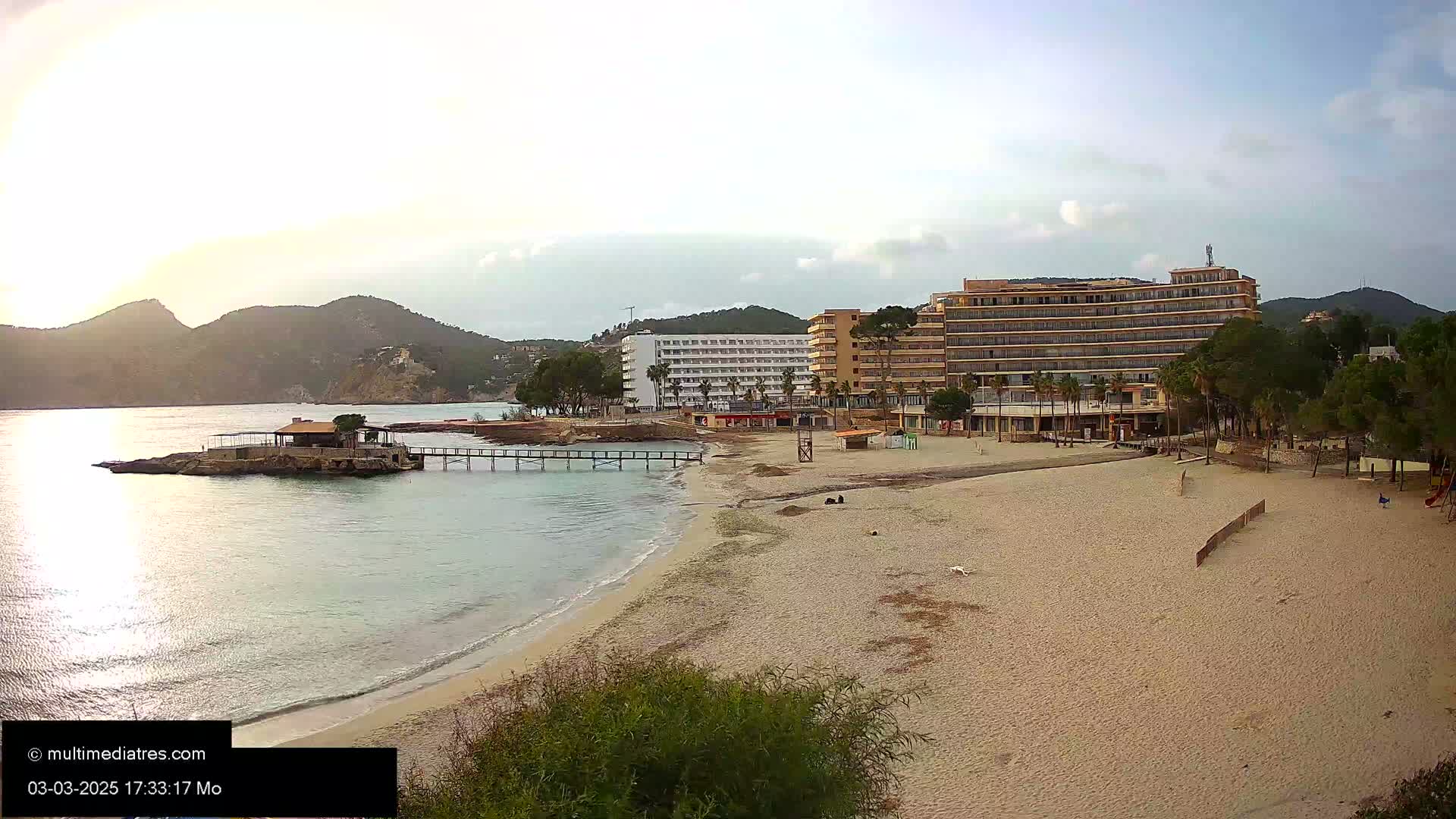 A sandy beach with calm, clear water laps the shore near a pier and several hotels, with mountains in the background under a mostly clear sky.
