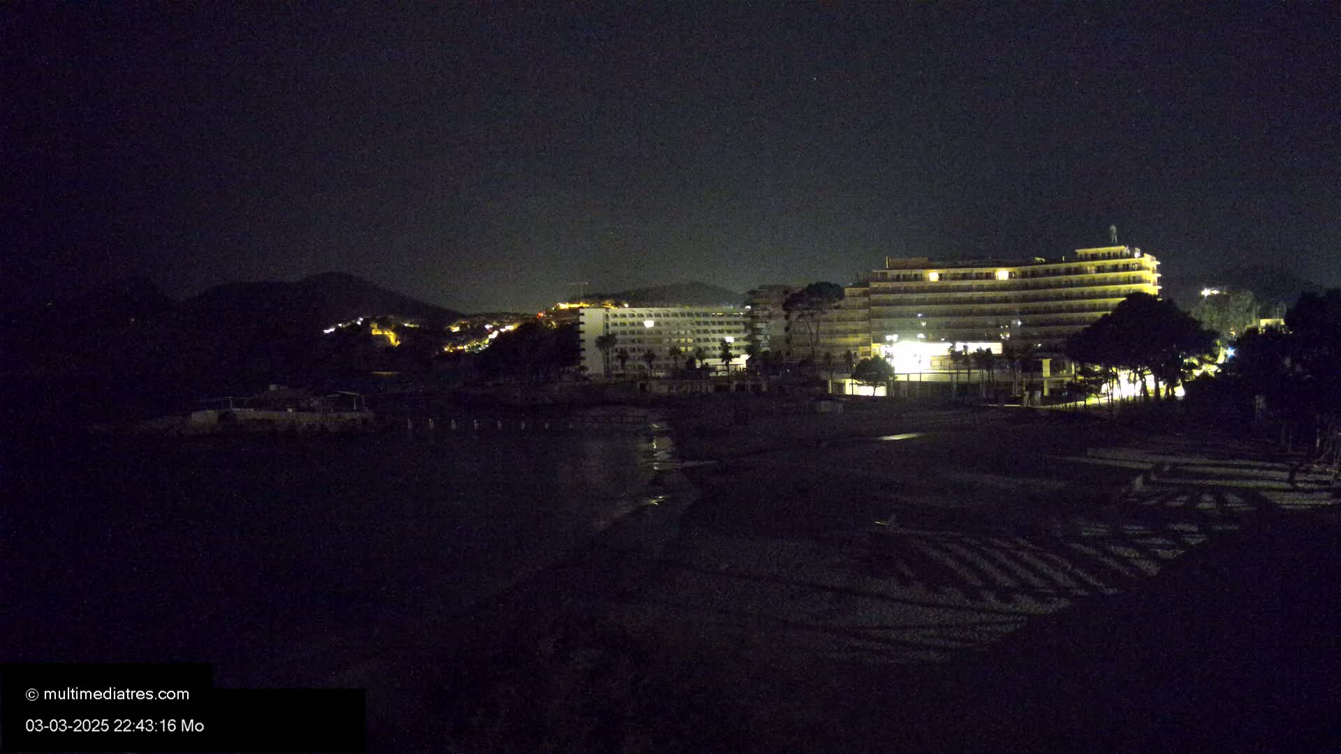At night, illuminated hotels and a hill with lights are visible across a dark beach.