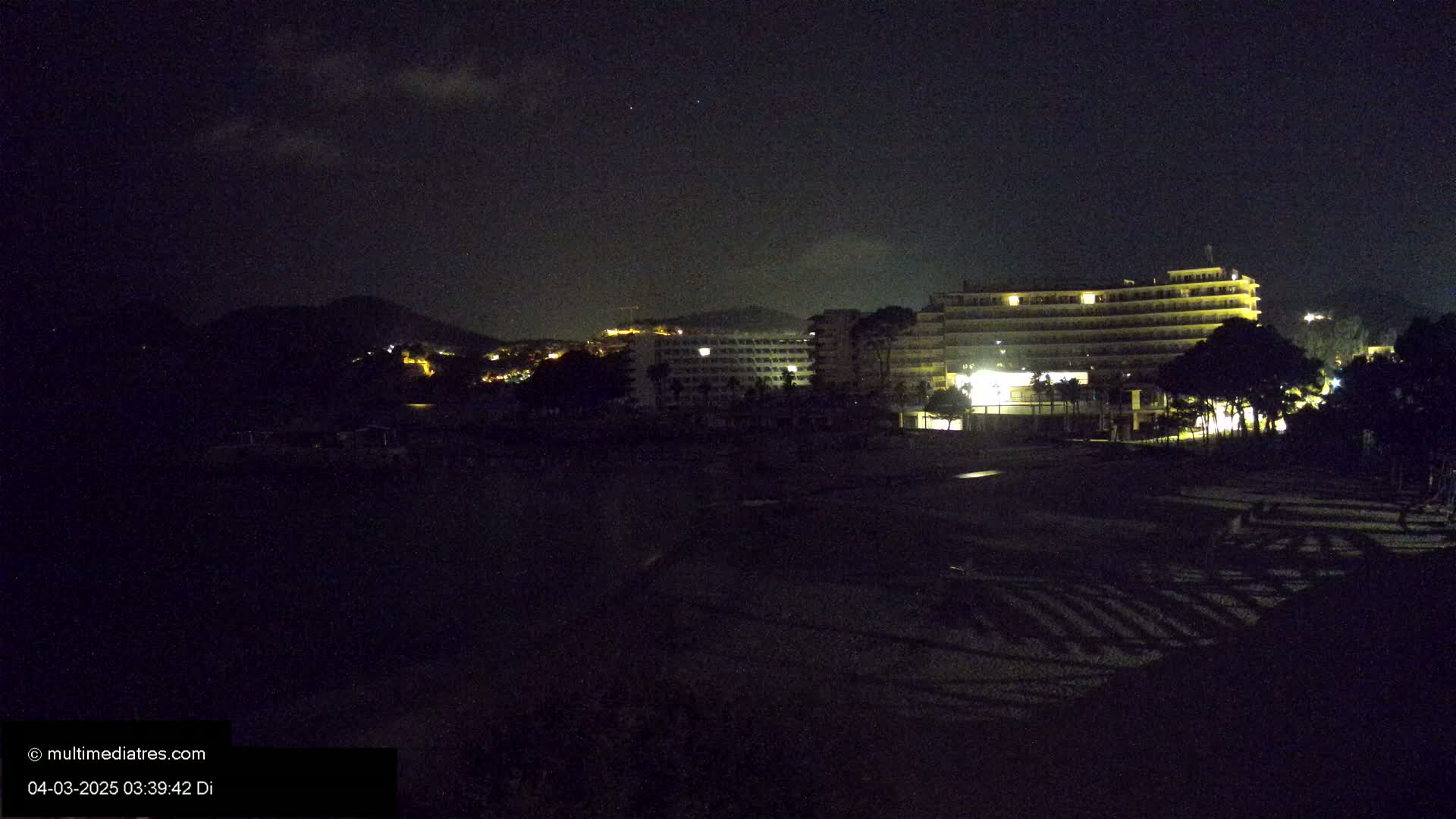 A nighttime view of a dark beach with illuminated hotels and a hill in the background under a partly cloudy sky.