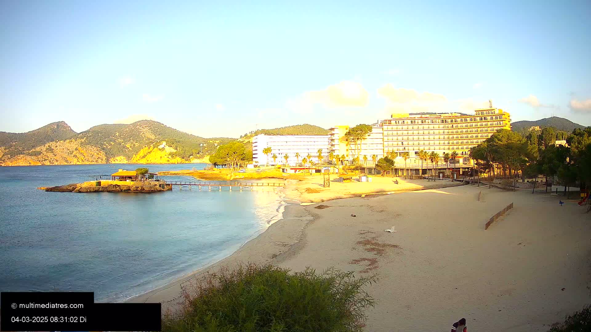 A sandy beach with a large hotel and a small island with a building are visible under a clear, bright sky near mountainous terrain.