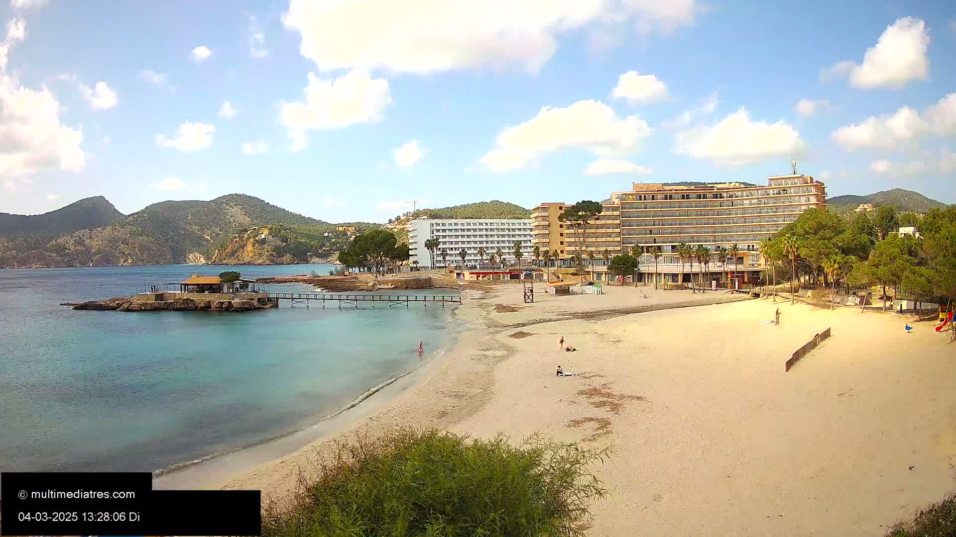 A sandy beach with a few people on it sits in front of hotels and green hills under a partly cloudy sky.