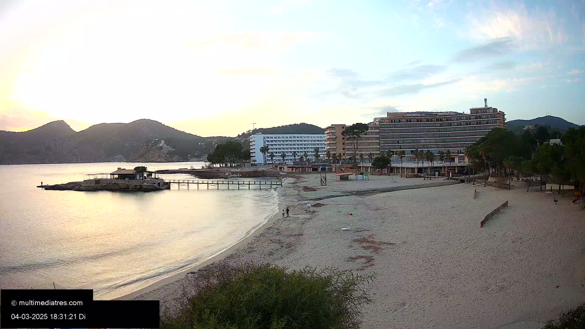 A sandy beach with a calm sea, a pier, and several hotels is seen at sunset, with mountains in the background.