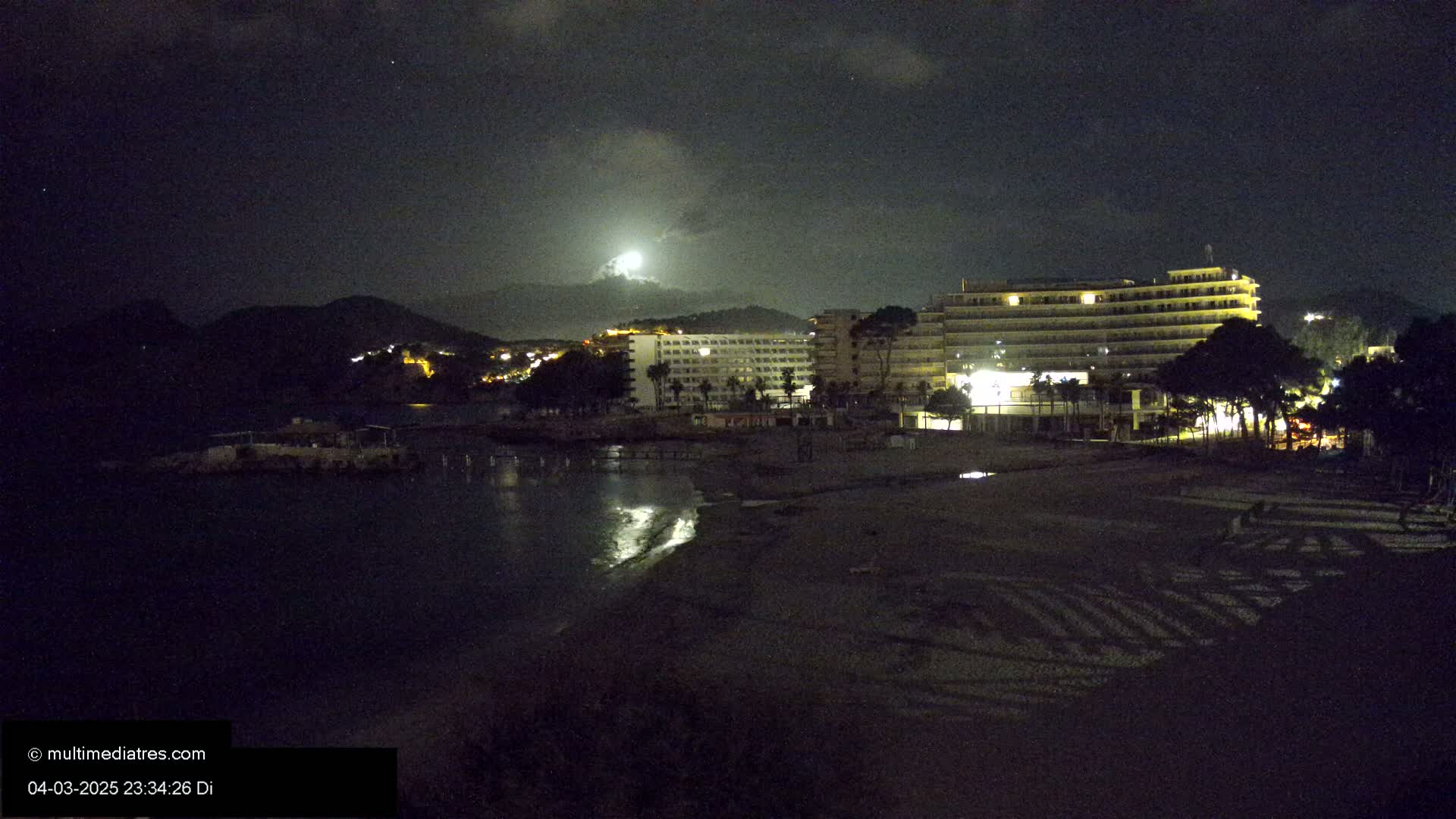 A moonlit nighttime scene shows a beach with a large hotel and other buildings, illuminated against a dark mountain backdrop under a cloudy sky.
