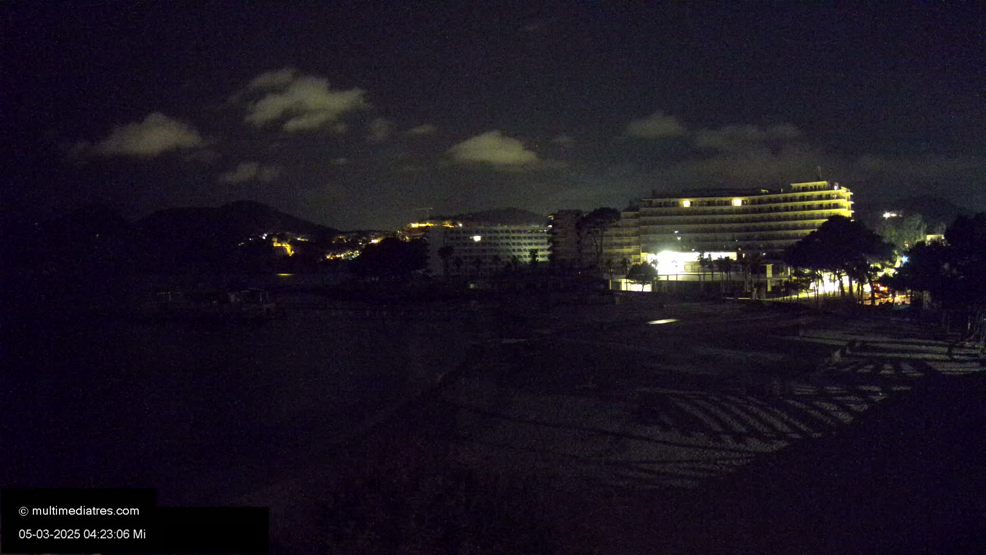 A nighttime view of a beach with illuminated hotels and a hill in the background under a partly cloudy sky.