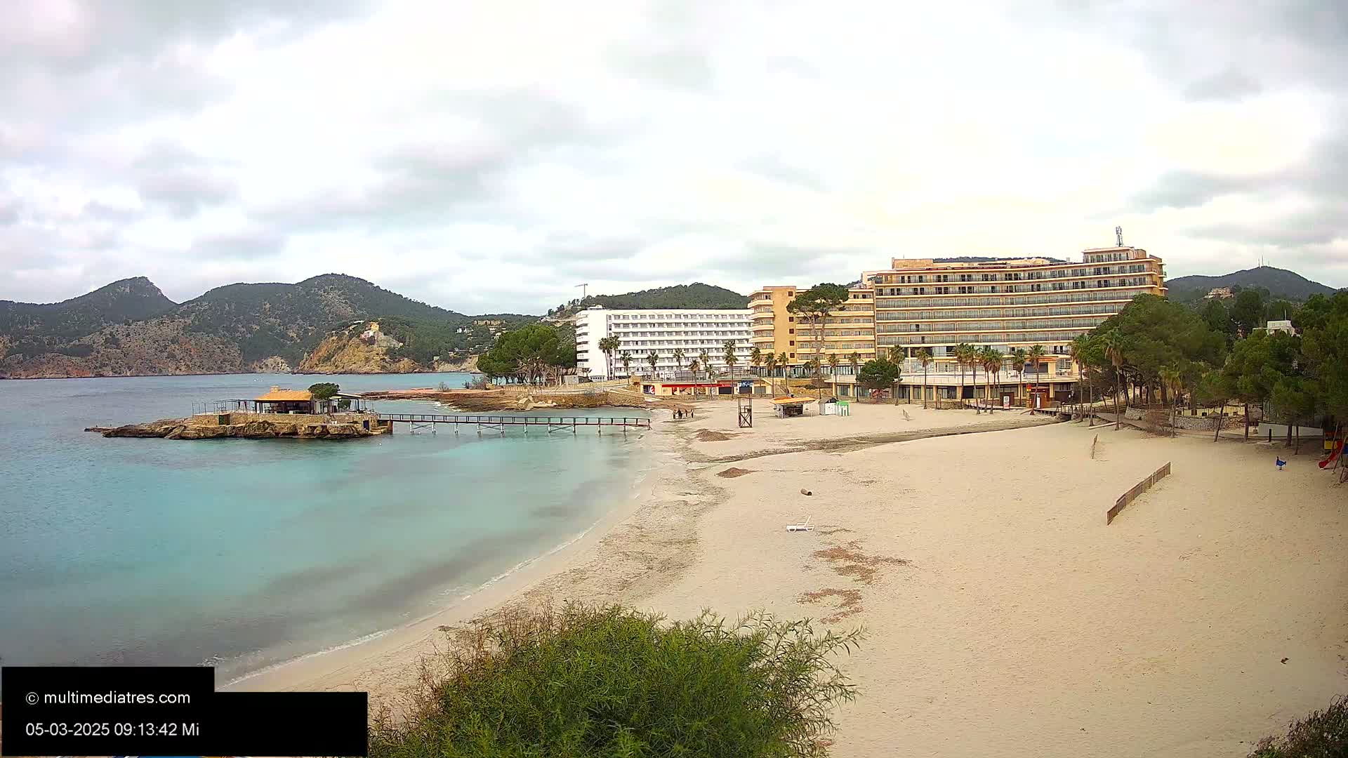 A sandy beach with turquoise water, a small pier, and several hotels sits beneath a cloudy sky against a backdrop of mountains.