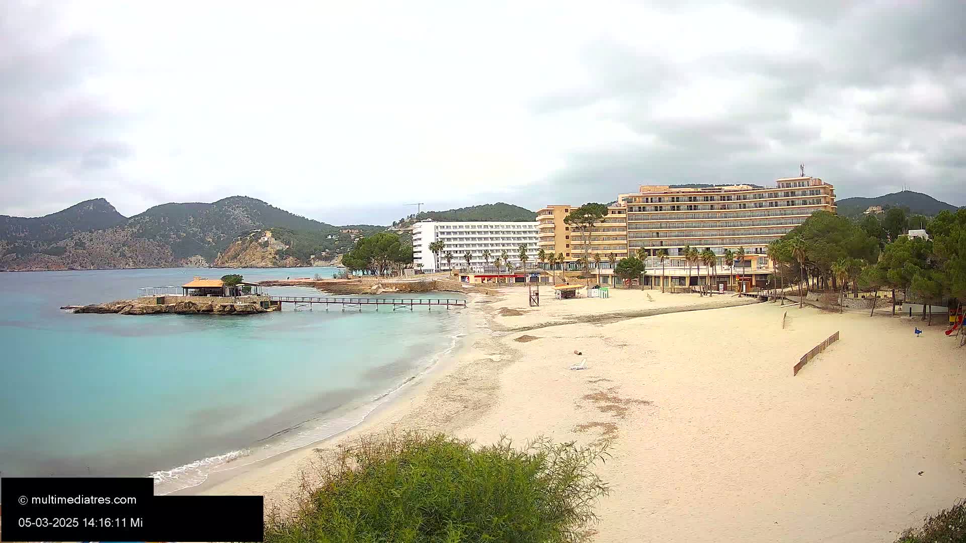 A sandy beach with turquoise water, a small beachfront building on a pier, and two large hotels behind it under an overcast sky.