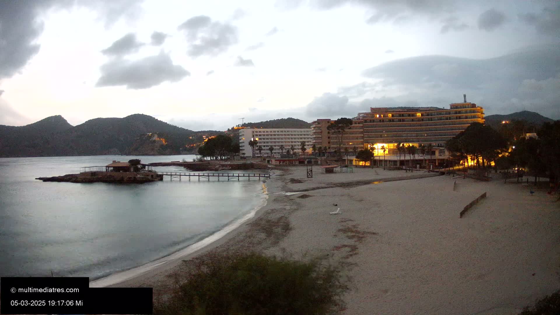 A sandy beach at dusk under a cloudy sky, featuring a small island with a building, a long pier, and several hotels illuminated along the shoreline.