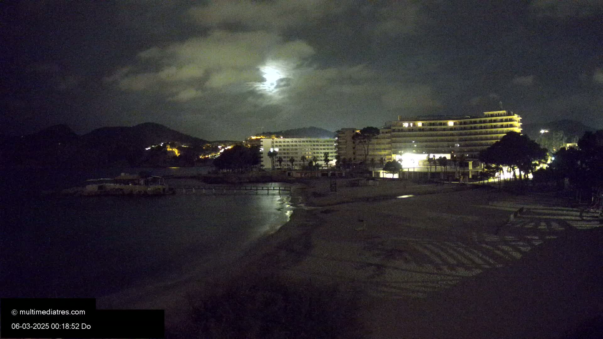 A moonlit night scene shows a dark beach with a large hotel and other buildings behind it, all illuminated, with a dark mountain in the background under a cloudy sky.