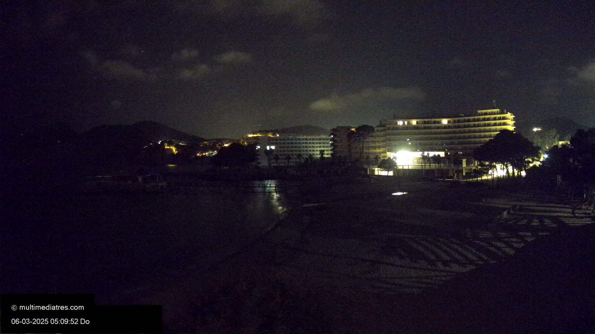 A nighttime view of a beach with several illuminated hotels and a hill in the background under a partly cloudy sky.