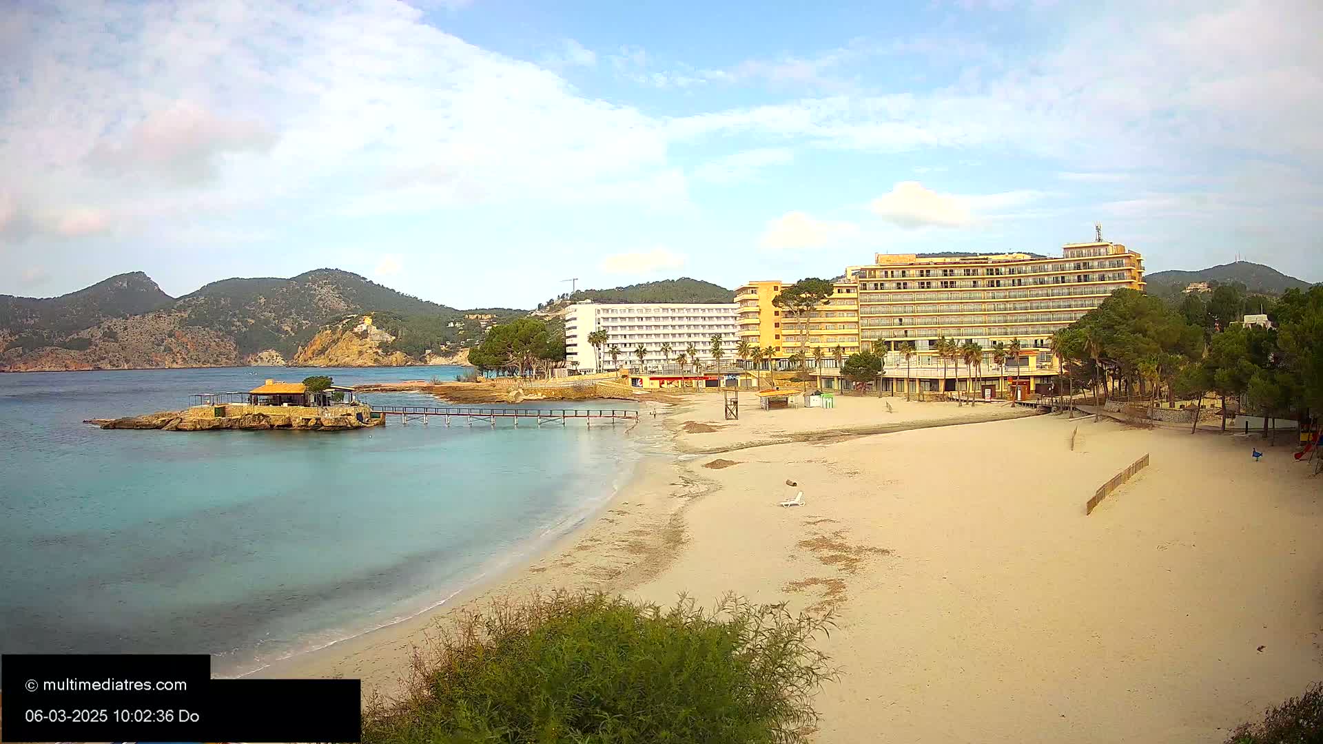 A sandy beach with calm, clear water, a pier, a small building on a rock outcropping, and large hotels in the background under partly cloudy skies.