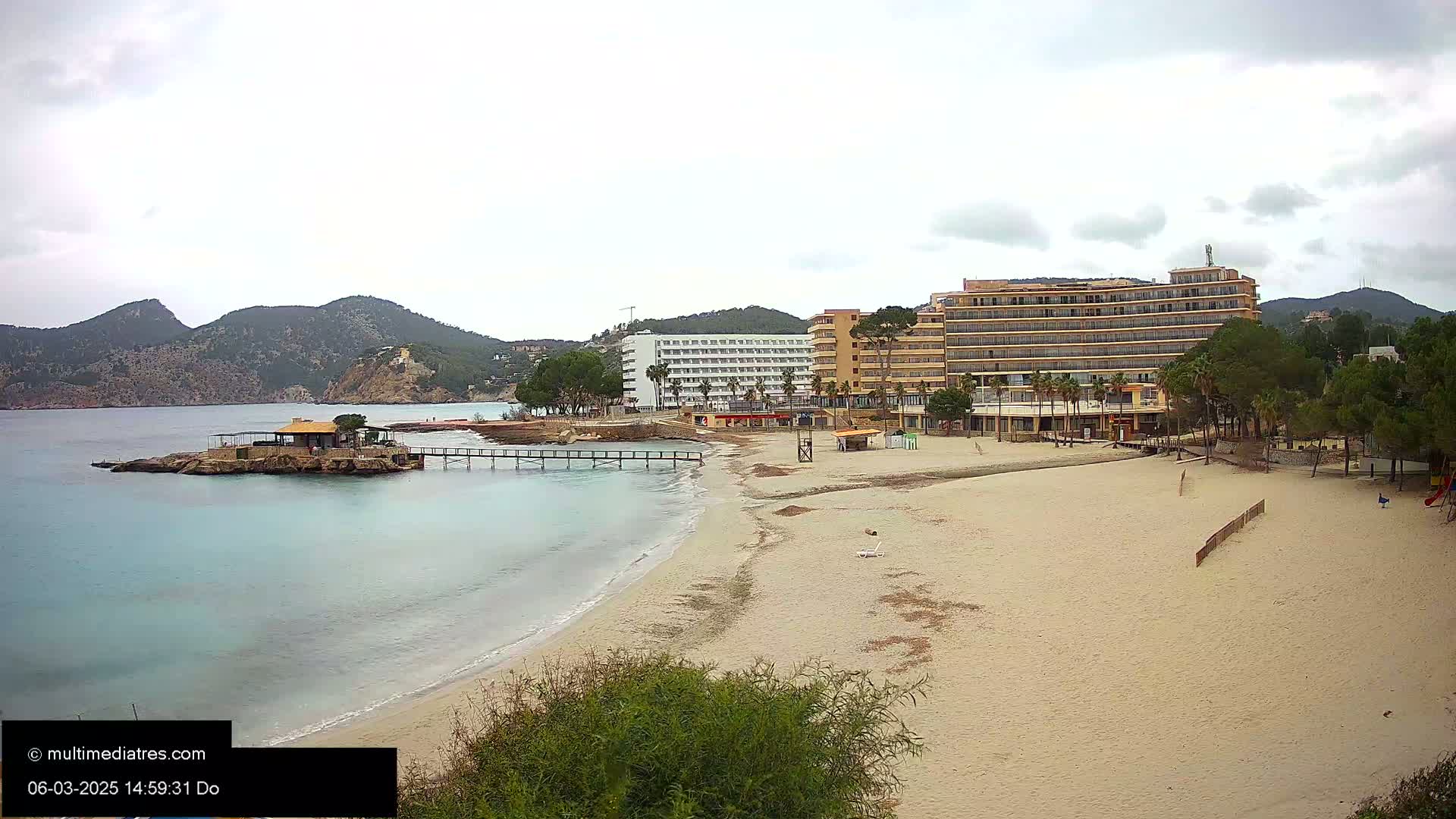 A sandy beach with calm, light-blue water stretches out toward two hotels and a small, rocky island with a building on it, all under an overcast sky.