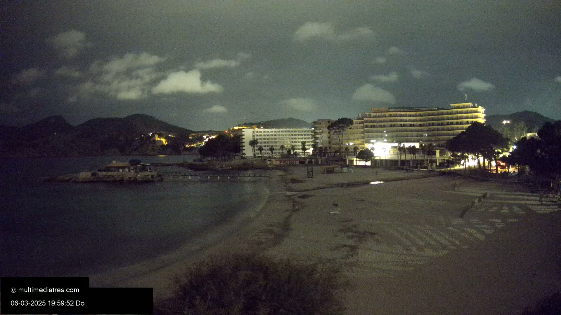 A mostly cloudy night scene shows a beach at night with a large hotel and other buildings in the background, near dark hills.