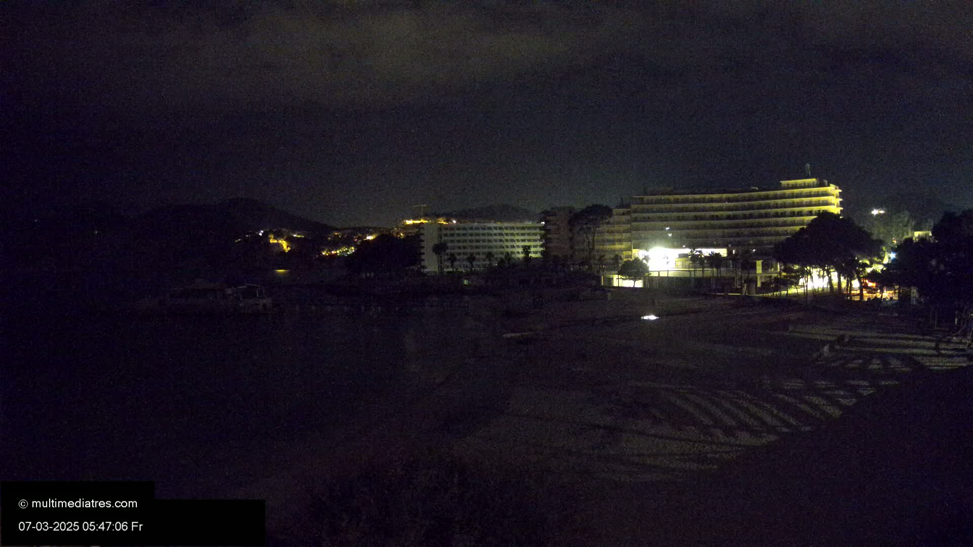 At night, a dimly lit beach is shown with several large buildings in the background under a dark, cloudy sky.