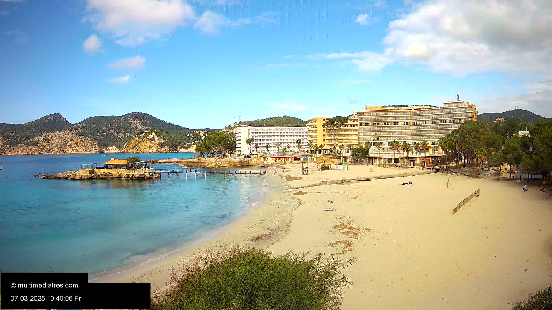 A sandy beach with calm, turquoise water stretches before two hotels, backed by low mountains under a mostly sunny sky with some clouds.