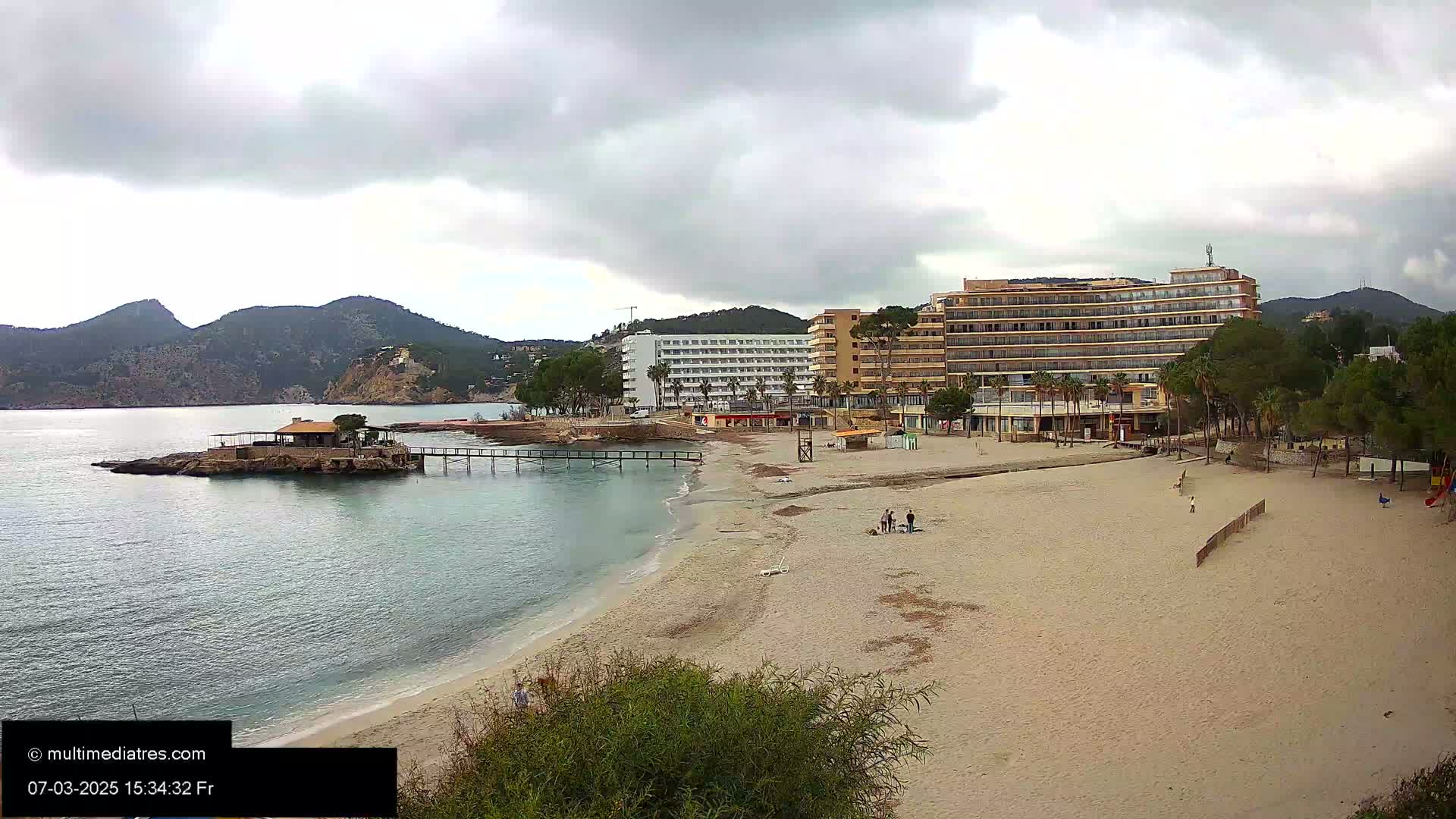 A sandy beach with a few people, hotels, and a small island with a structure on it is visible under an overcast sky.