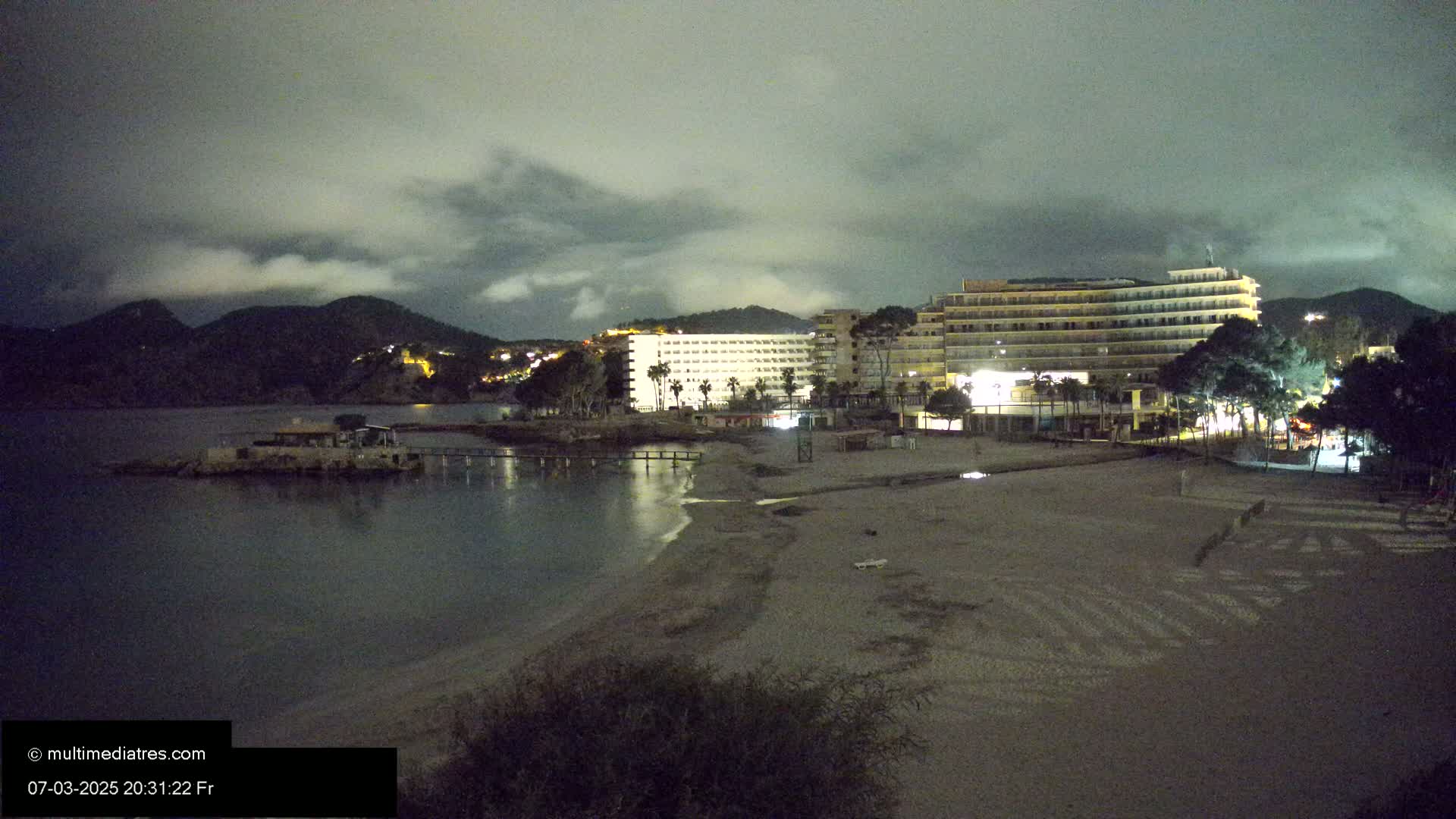 A nighttime view of a beach with several lit buildings and palm trees behind it, under a cloudy sky.