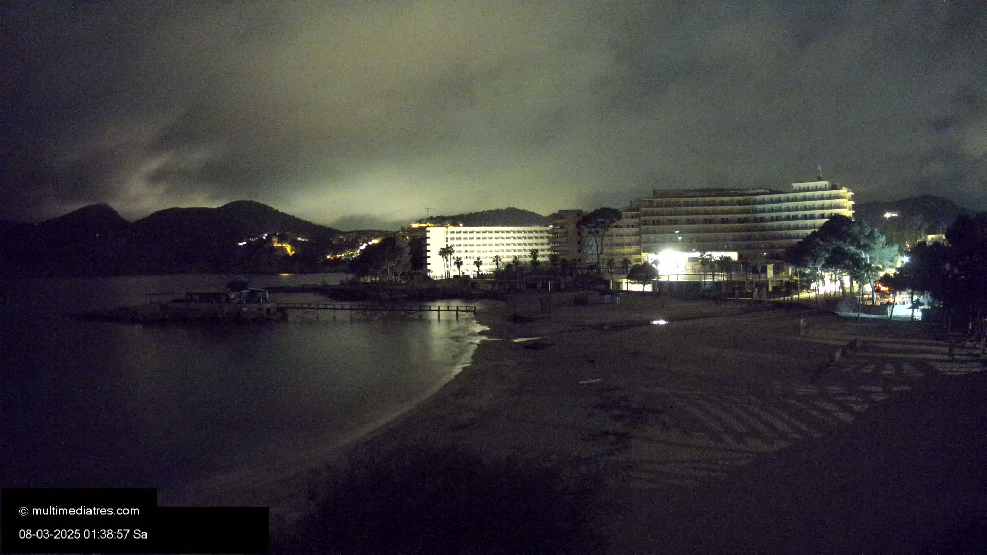 A dark, nighttime beach scene shows a calm body of water, a sandy shoreline with a crosswalk pattern visible, and several illuminated hotels against a backdrop of dark mountains under an overcast sky.
