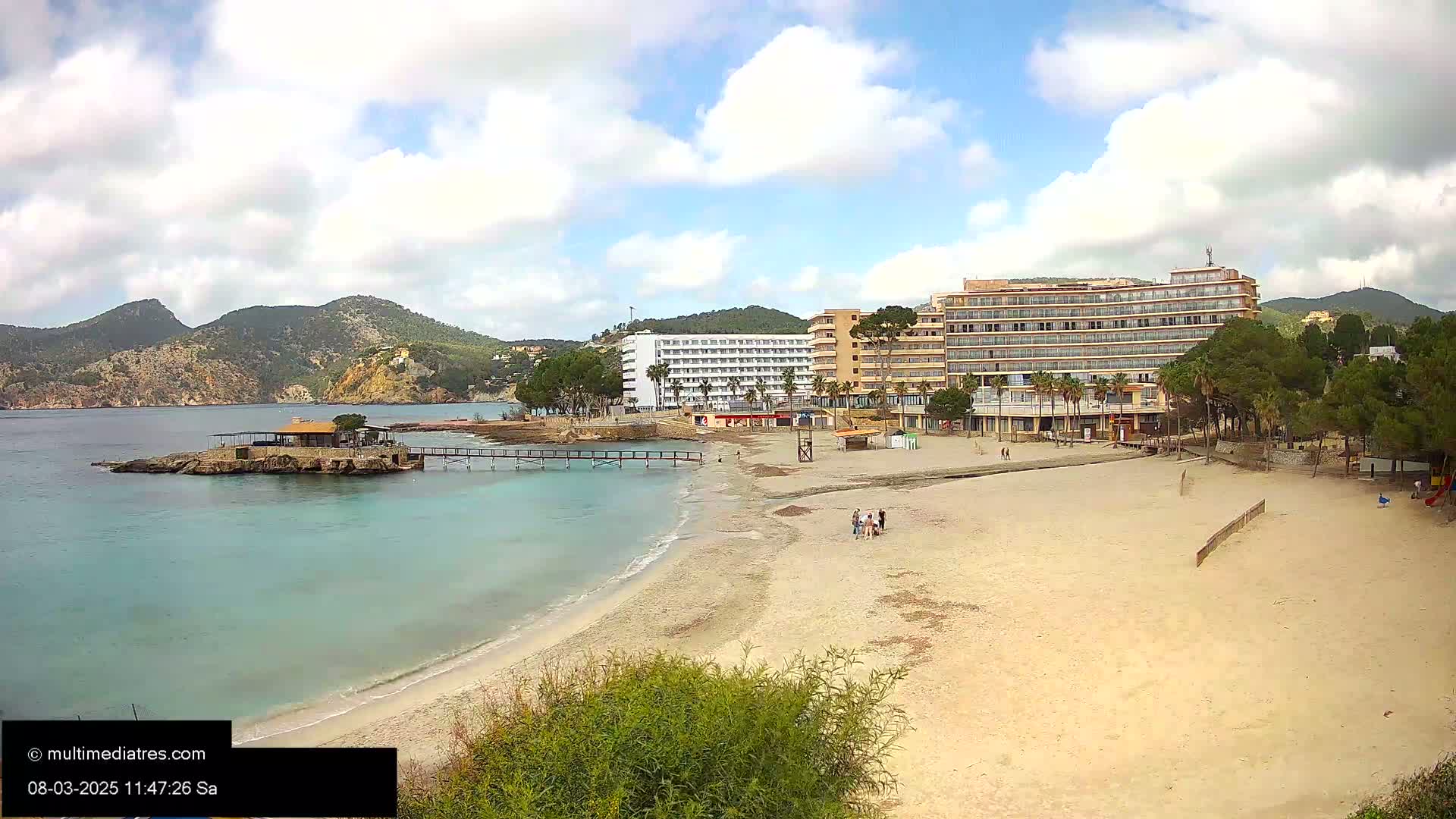 A sandy beach with a few people on it sits below two large hotels, a small island with a building, and a rocky, mountainous coastline under a partly cloudy sky.