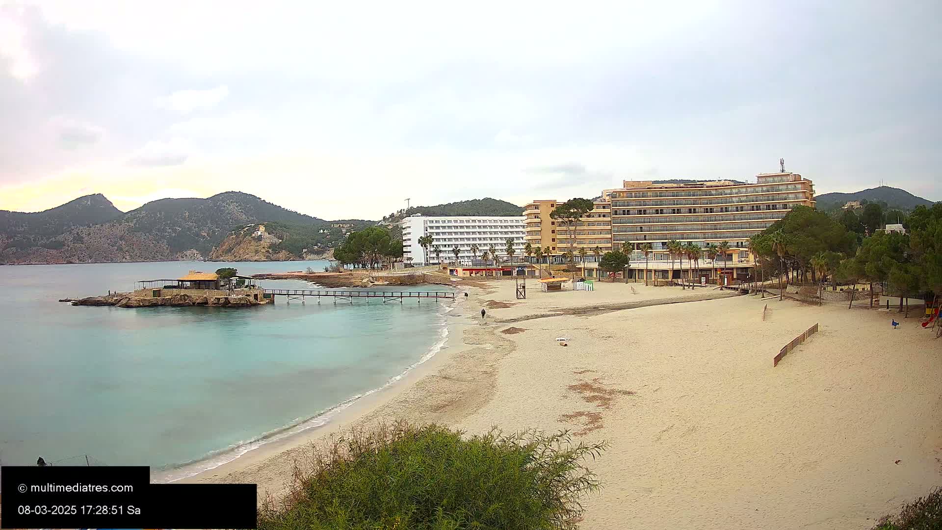 A sandy beach with calm, turquoise water stretches out in front of two large hotels and a small island with a building on it, all under an overcast sky.