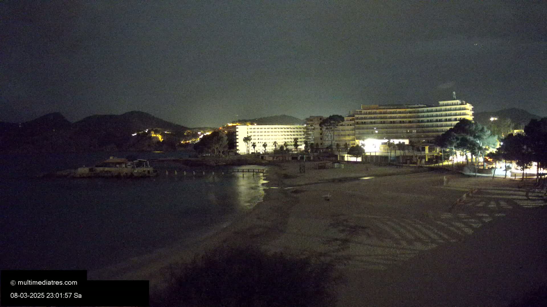 At night, a dark sandy beach is bordered by lit hotels and a calm body of water under a cloudy sky, with hills visible in the distance.