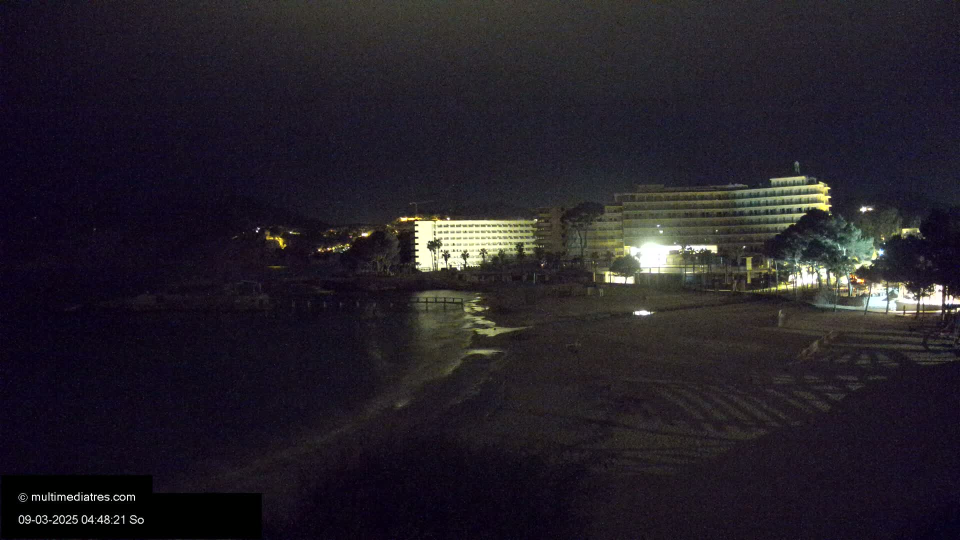 A dark night scene shows a beach and a large, brightly lit hotel building near the water.