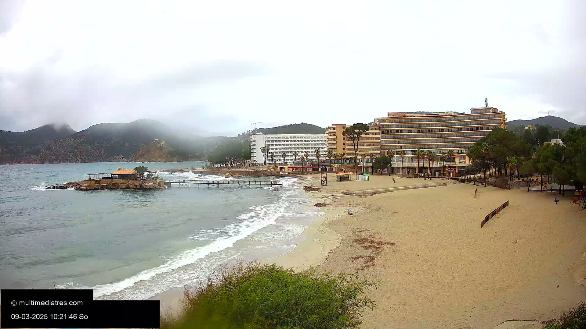 A sandy beach with a small jetty, hotels, and a restaurant on a rocky island, all under an overcast sky.