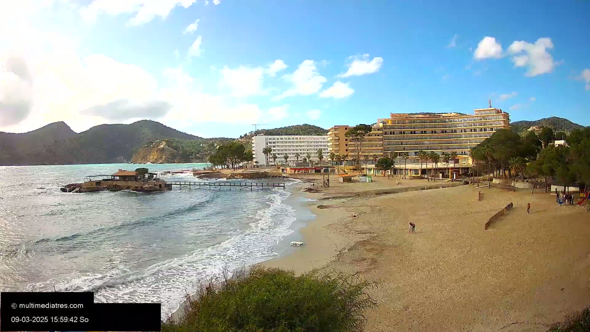A sandy beach with several hotels and palm trees behind it is partially surrounded by calm, wavy water, with mountains in the background under a partly cloudy sky.