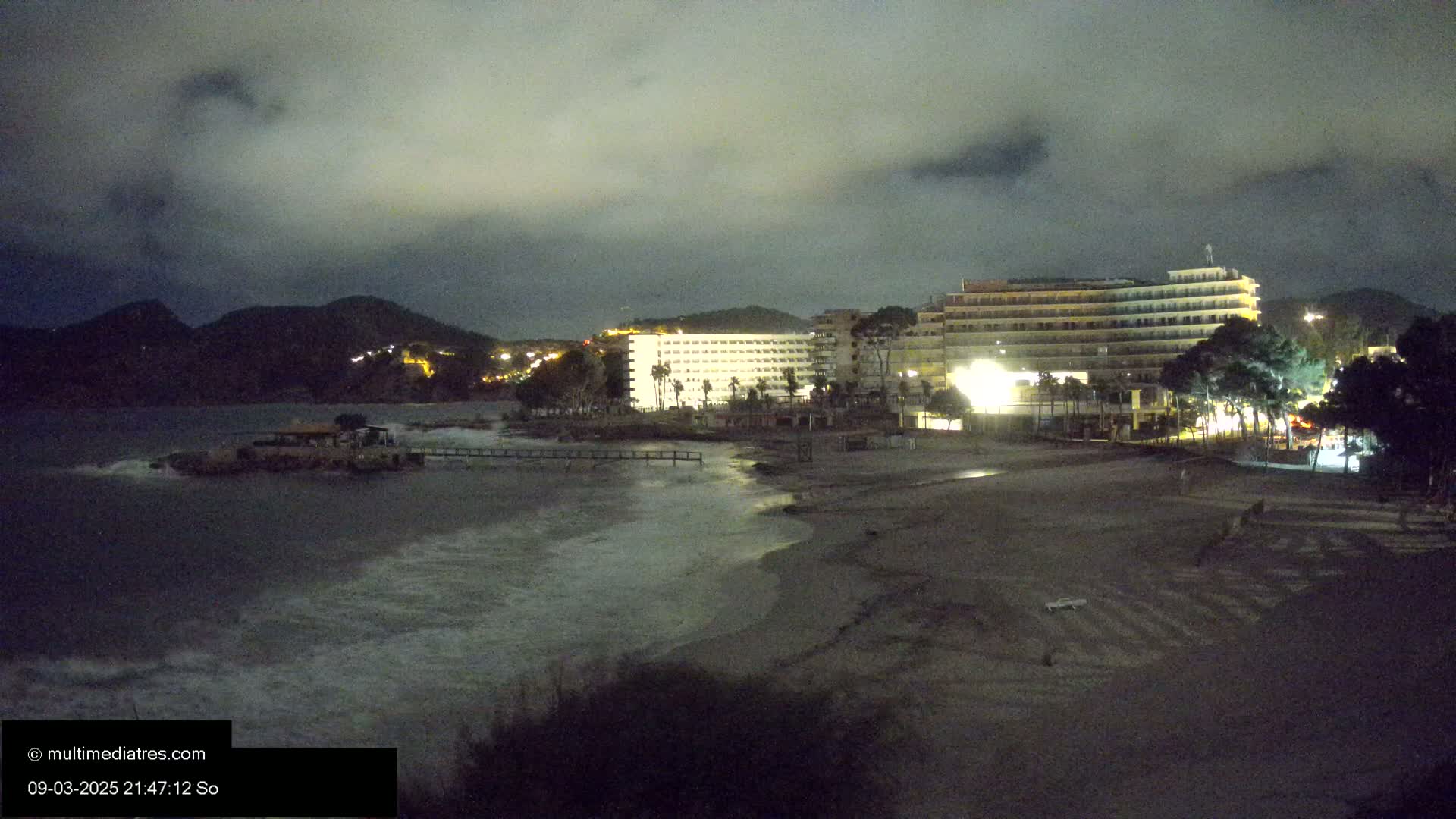At night, under a cloudy sky, a beach with a few buildings, some palm trees, and waves crashing on the shore is visible.