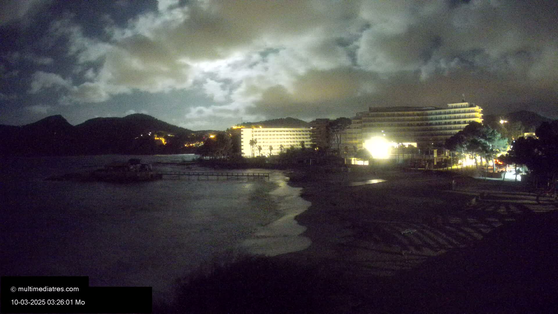 A dark beach at night, under a cloudy sky, with a brightly lit hotel and hills visible in the background.