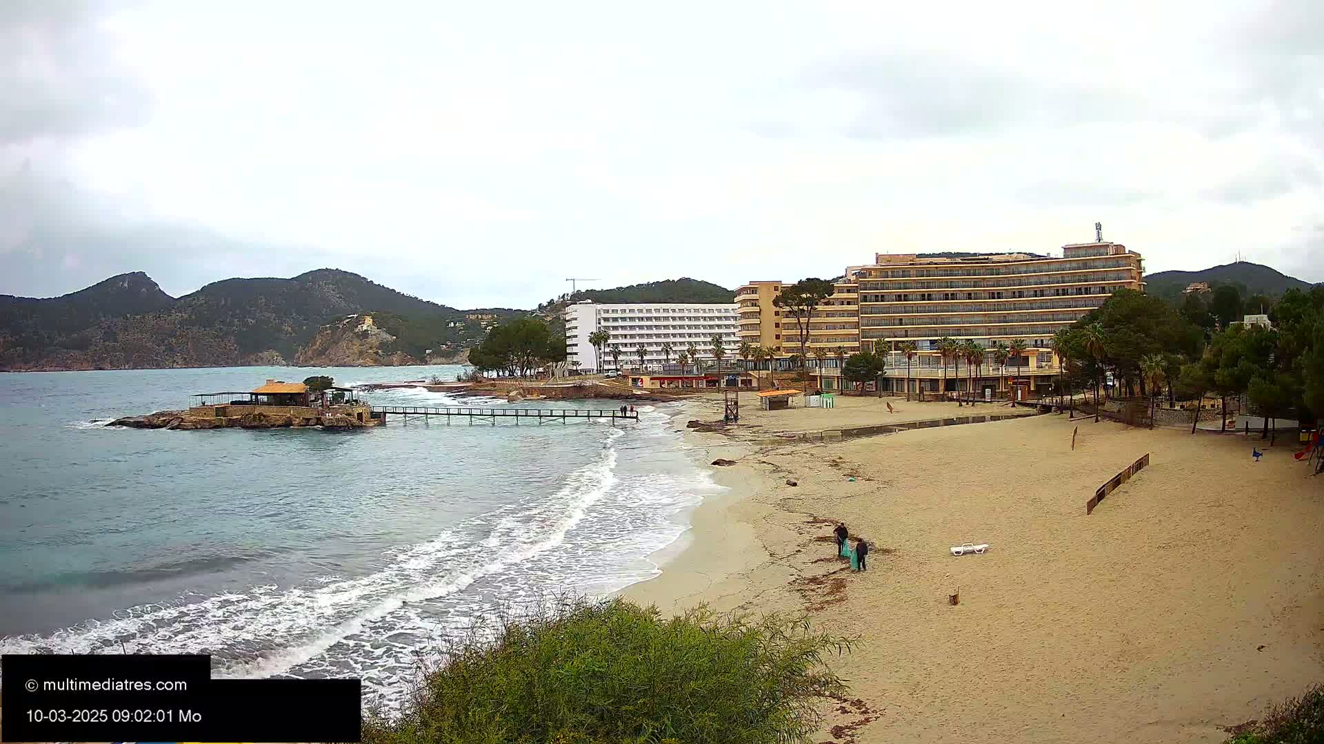 A sandy beach with gentle waves laps against the shore near hotels and a small island with a building on it under an overcast sky.