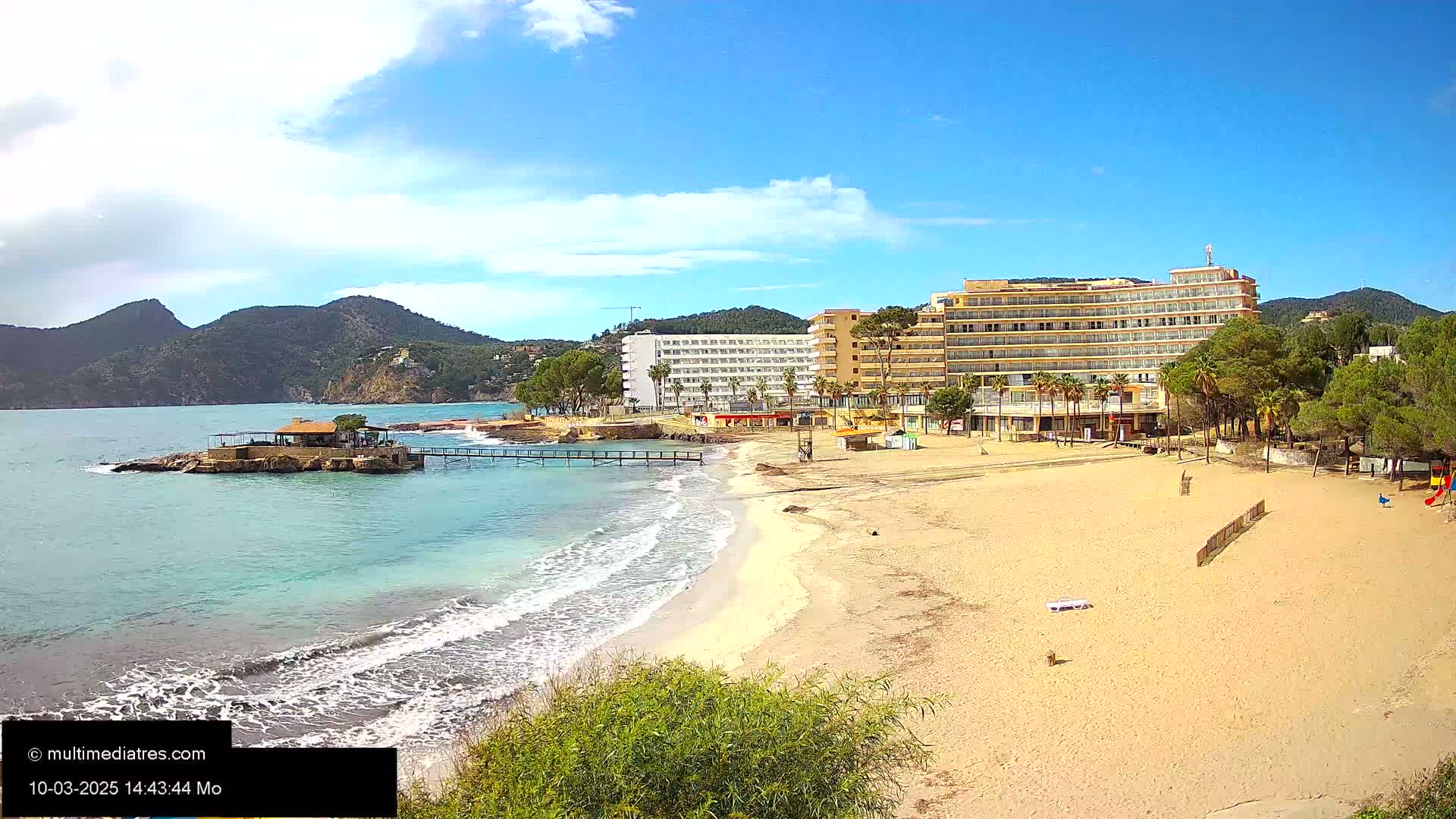 A sandy beach with a small pier and large hotel is seen from an elevated vantage point on a partly cloudy day.