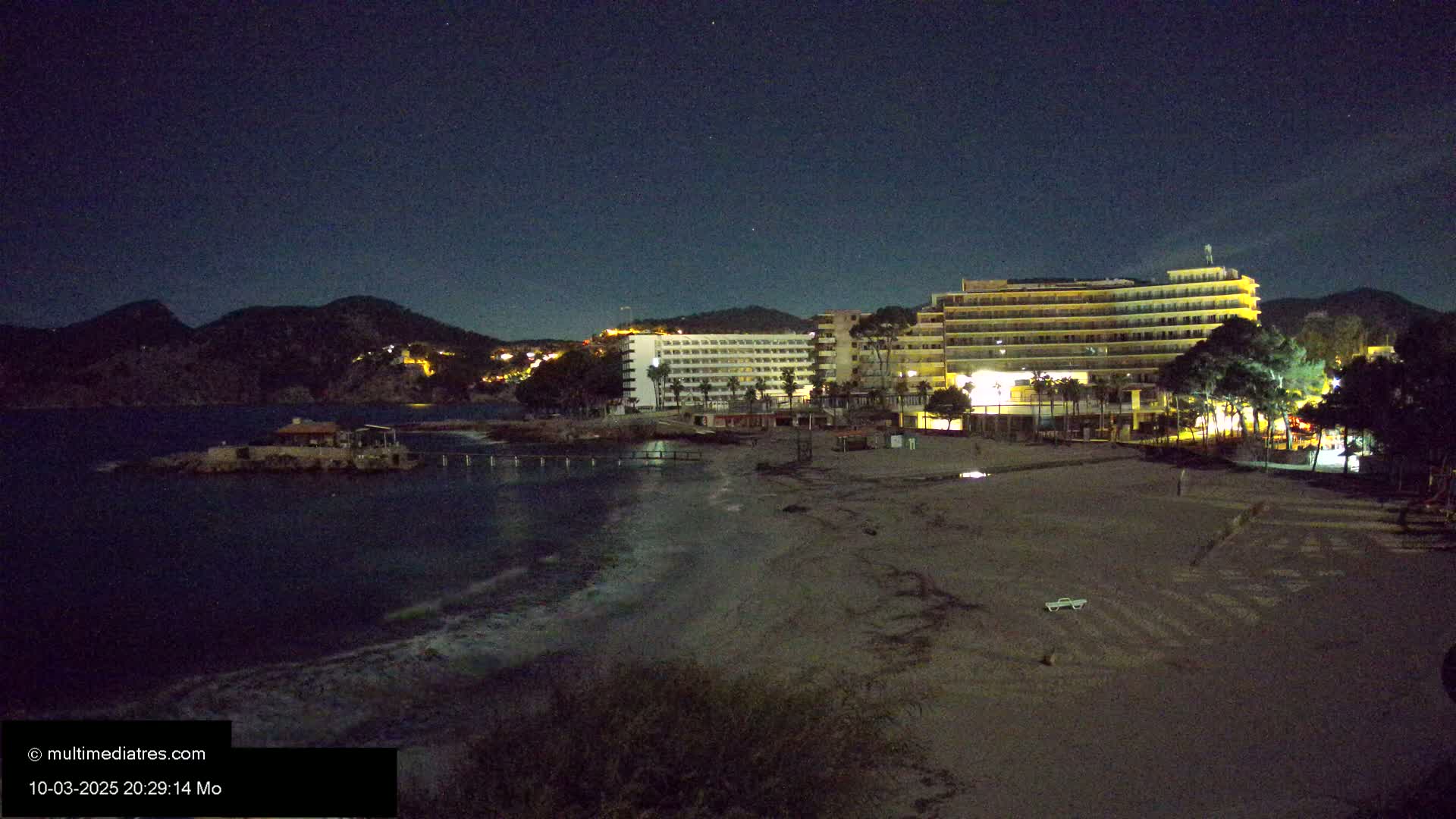 A dark, night-time view of a sandy beach with illuminated hotels and buildings on a hillside overlooking a calm body of water under a starry sky.