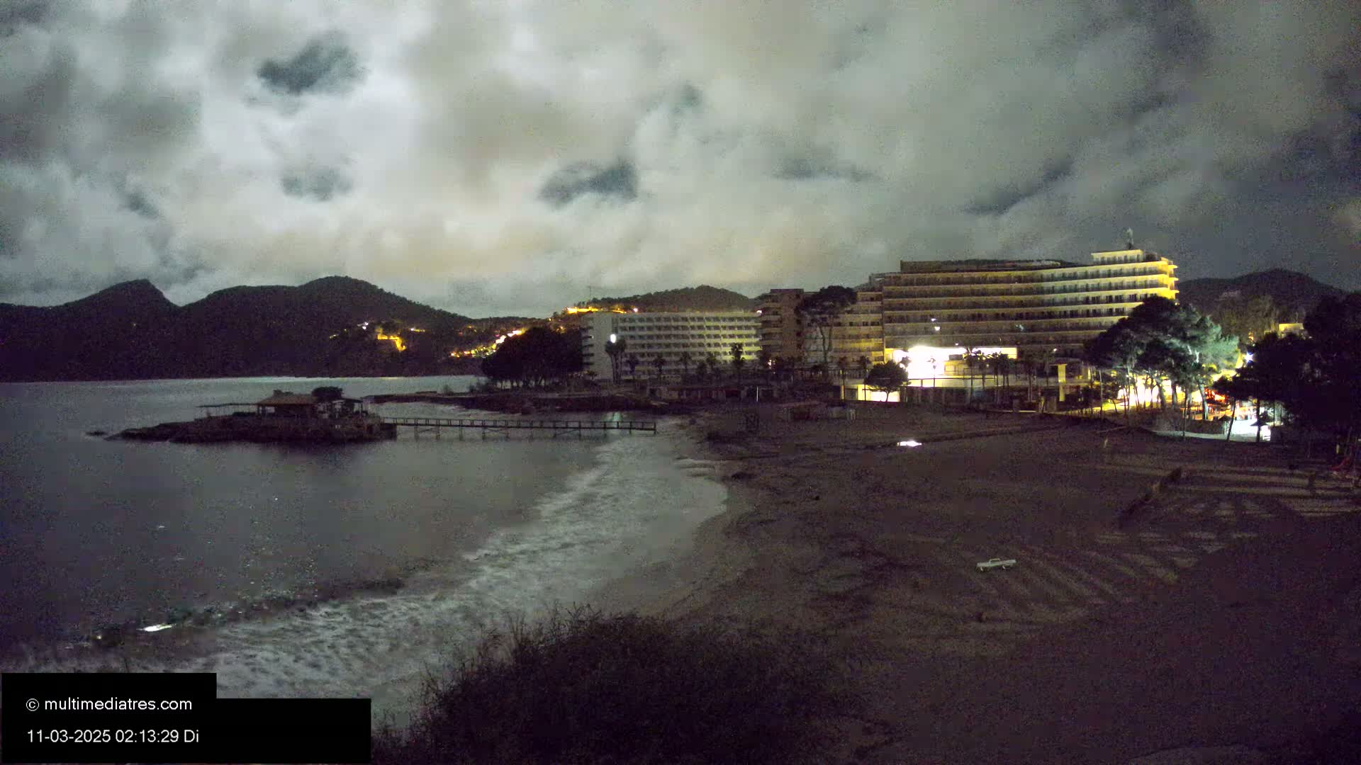 At night, under a cloudy sky, a dark beach with a few lights from buildings and some small waves laps against the shore, next to a dark body of water and some lit buildings.