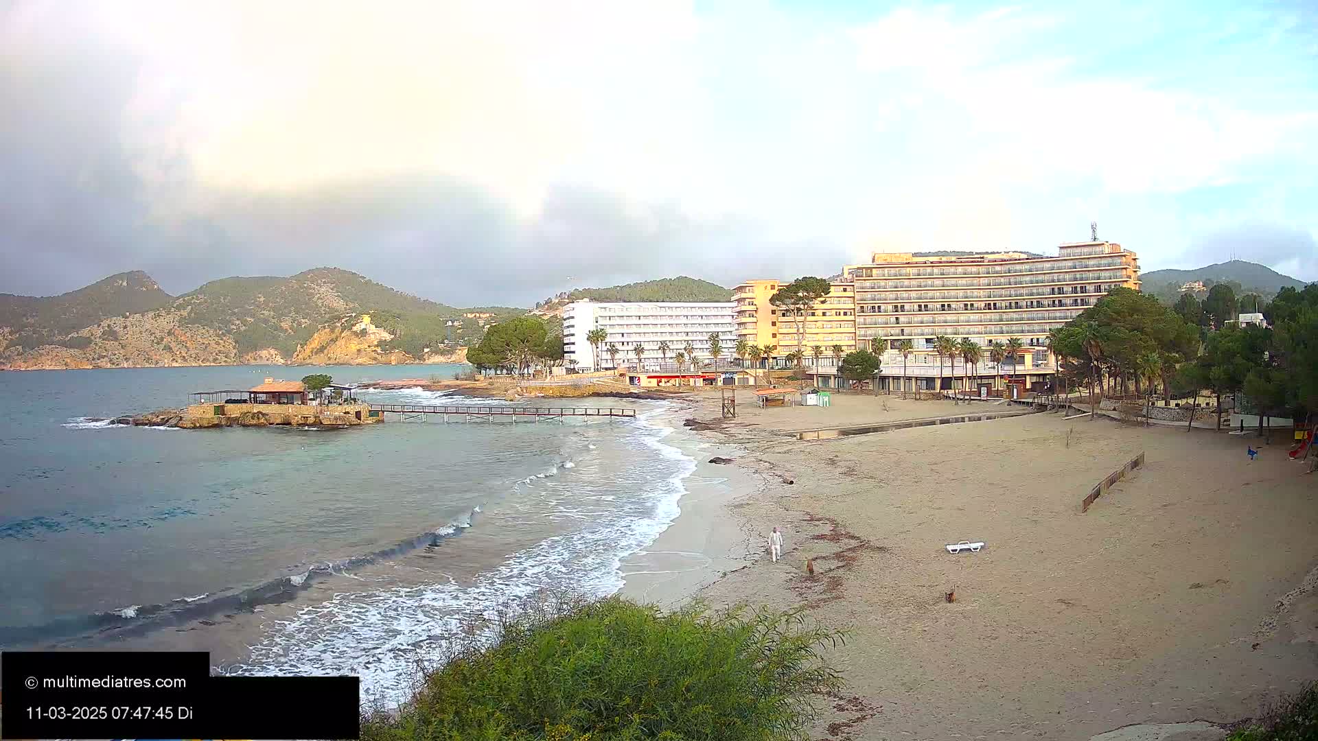 A sandy beach with small waves laps at the shore near several hotels and a small island with a building under a partly cloudy sky.