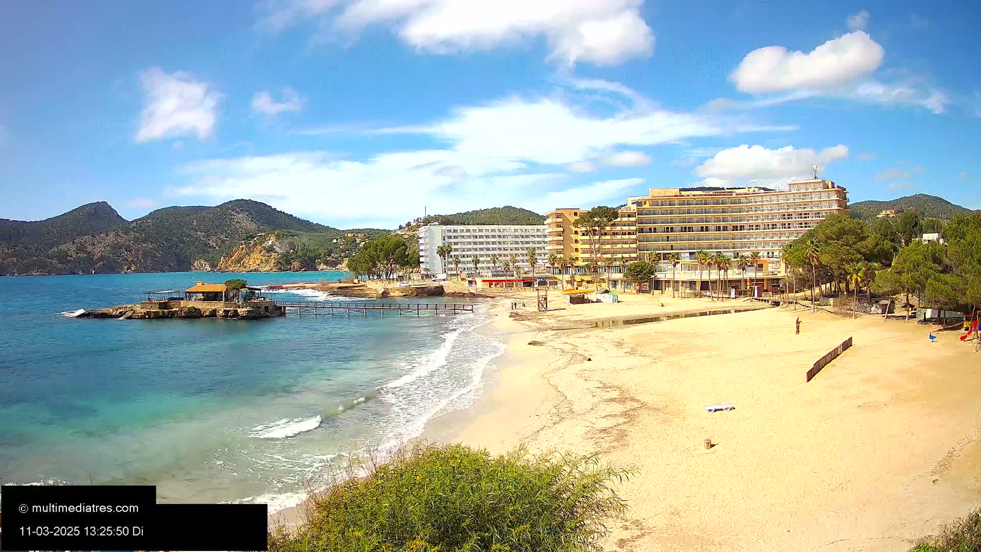 A sandy beach with gentle waves laps the shore, under a partly sunny sky, next to a row of hotels and lush green hills.