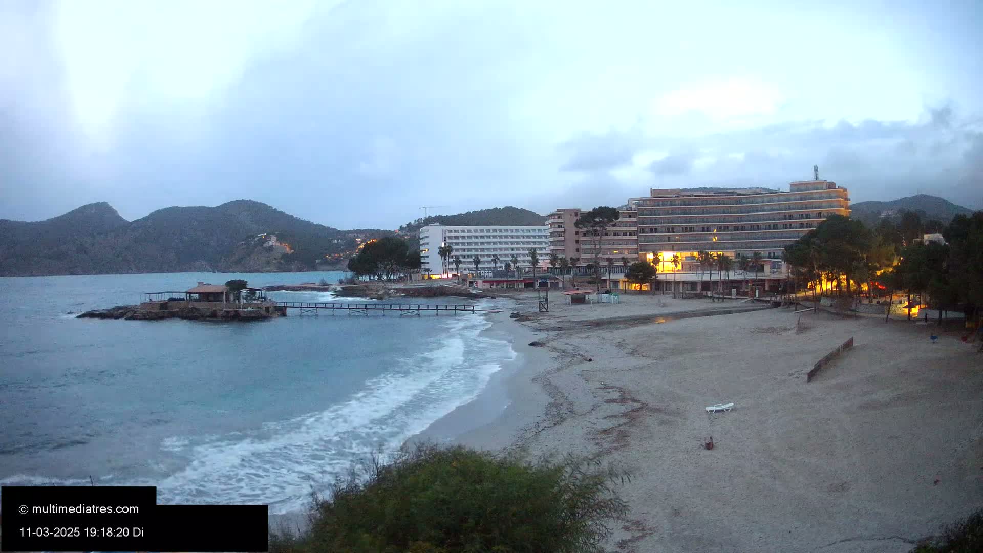 A sandy beach at twilight, with small waves lapping the shore, overlooks a calm bay with a small island and several hotels under a cloudy sky.