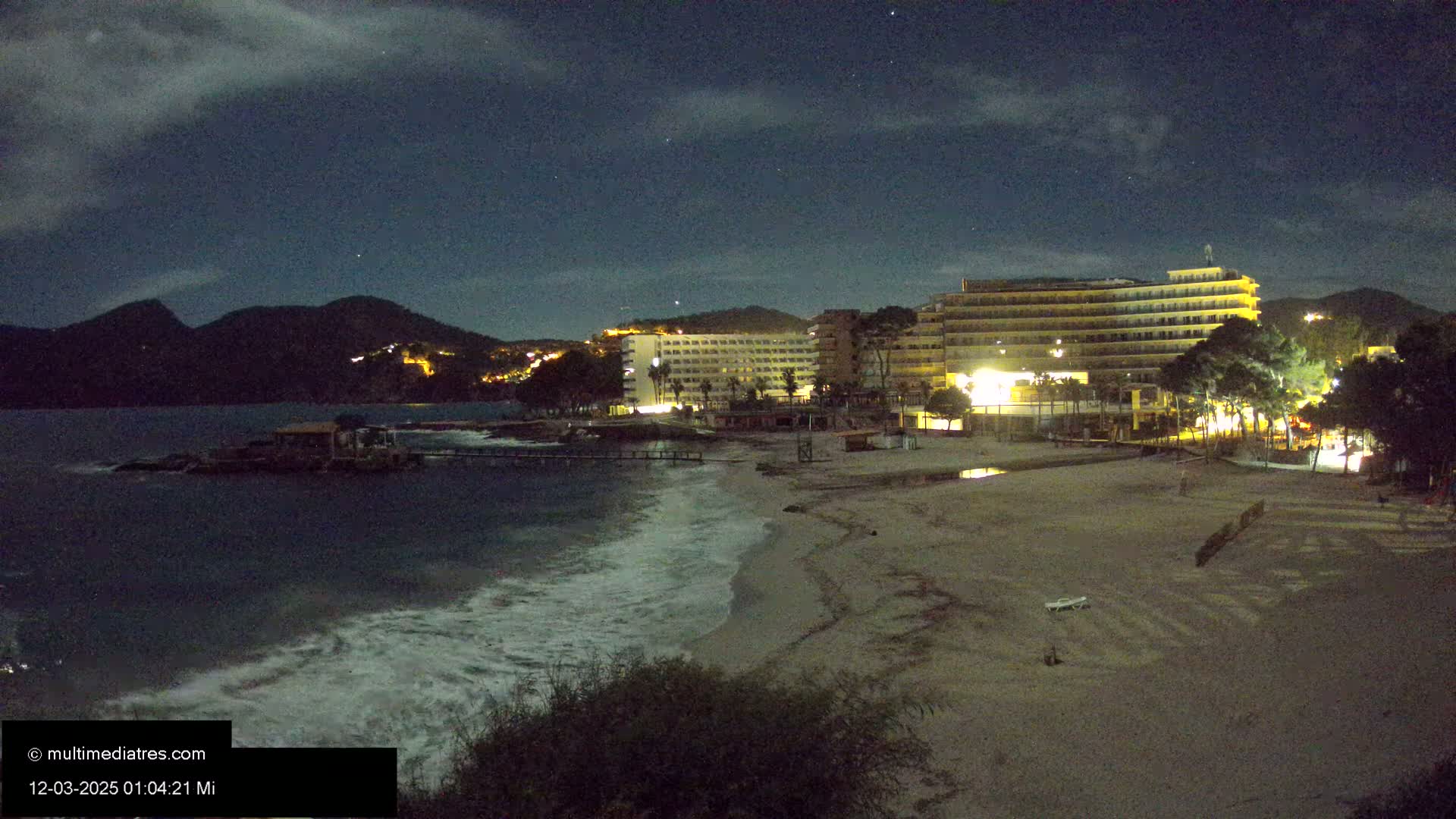 Under a partly cloudy night sky, waves lap a sandy beach in front of illuminated hotels and a dark mountain range.