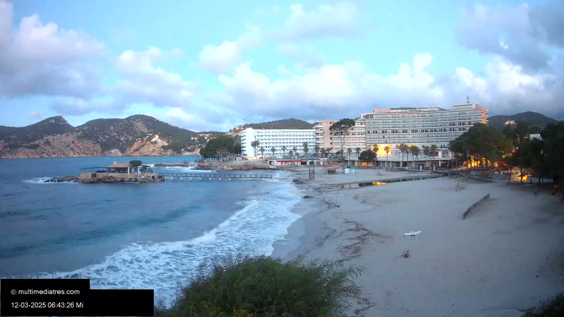 A sandy beach with small waves laps the shore near two large hotels under a partly cloudy sky.