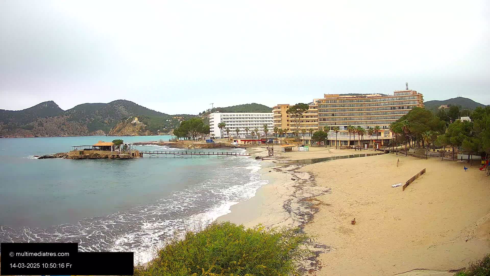A sandy beach with small waves laps the shore near several hotels and a small island with a building, all under an overcast sky.