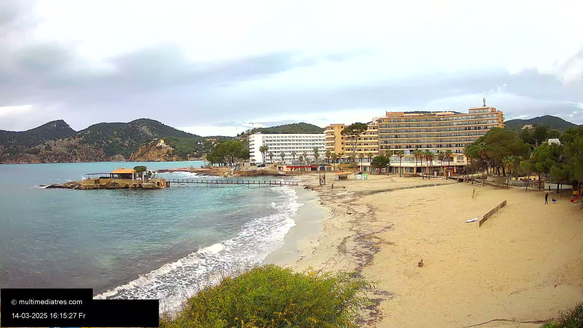 A sandy beach with calm waves laps the shore near a row of hotels, backed by lush green hills under a cloudy sky.