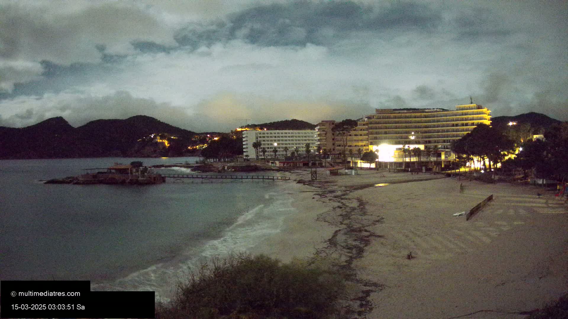 A dimly lit beach at night, under a cloudy sky, shows hotels and a small pier near dark mountains.