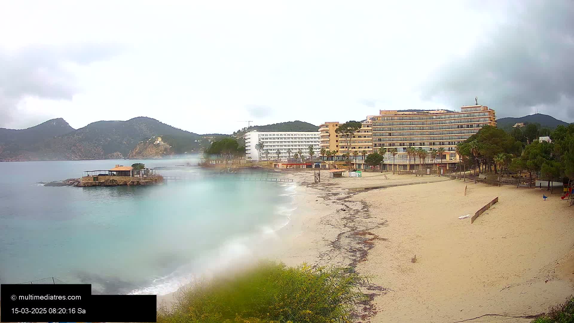 A sandy beach with hotels and palm trees is visible, alongside a calm, light-blue ocean under an overcast sky.